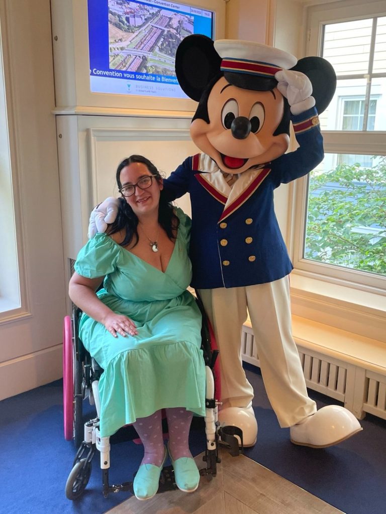 An image of a brunette wheelchair user in a turquoise dress seated next to mickey mouse in a sailor outfit