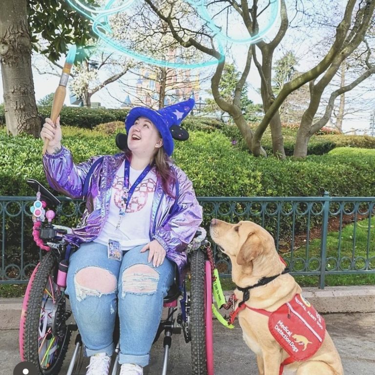 A magic shot featuring Lizzy seated next to her assistance dog Henry, painting mickey ears with a giant paintbrush