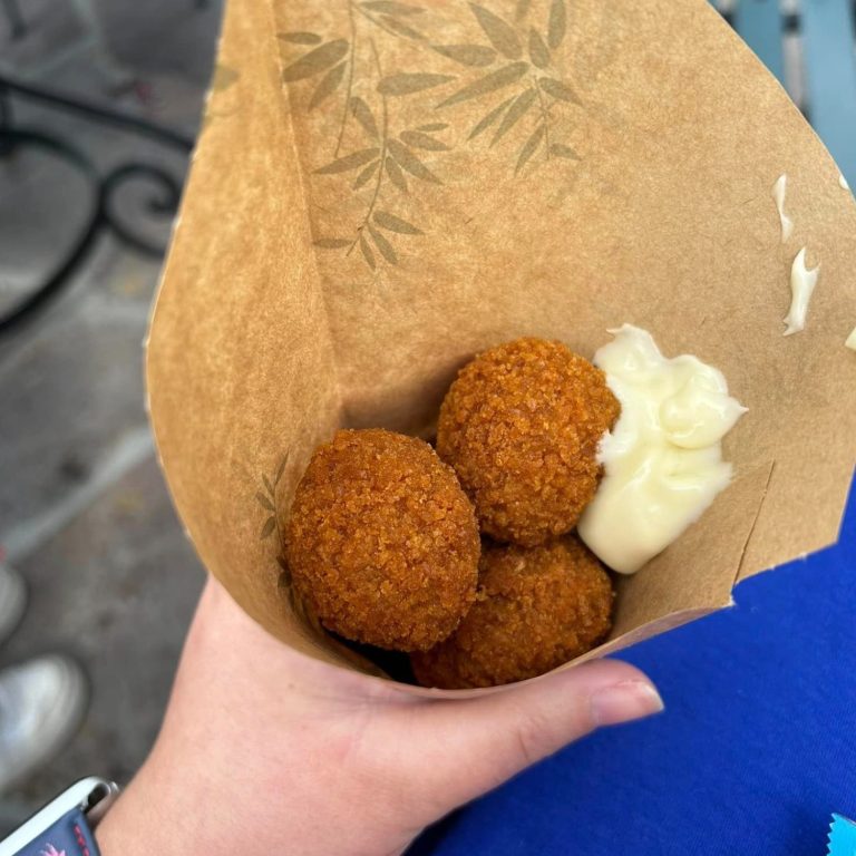 The image shows some round fried bitterballen snacks in a paper cone with some mayonnaise topping