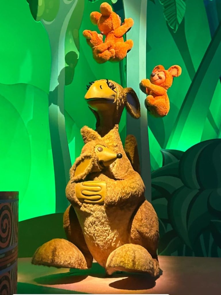 Image of a scene from it's a small world with a green background and a kangaroo with a loey with a tree behind it with Koala bears climbing it