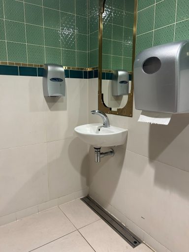 Tiled toilet cubicle with green and white tiles, a white sink and a hand towel dispenser