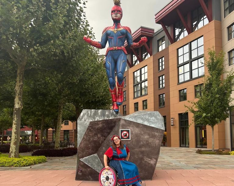 Image shows a captain marvel statue and in front of it is a wheelchair user dressed in a dress of a similar style to captain marvel