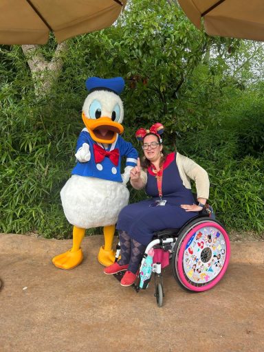 Image shows a female brunette wheelchair user in a blue dress seated next to donald duck, holding out his finger to indicate he is number 1