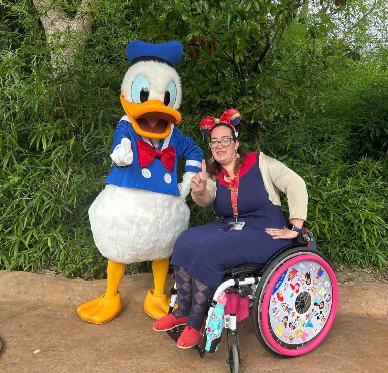 Jody is seated in her chair in a navy dress and holding her finger up like a number 1, next to donald duck who is holding his finger in the same way