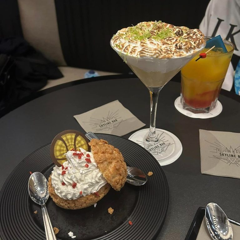 The image shows a cocktail in a martini glass which is yellow and has meringue topping, next to a choux bun filled with cream with a chocolate decoration which looks like the window in the Dr strange movies