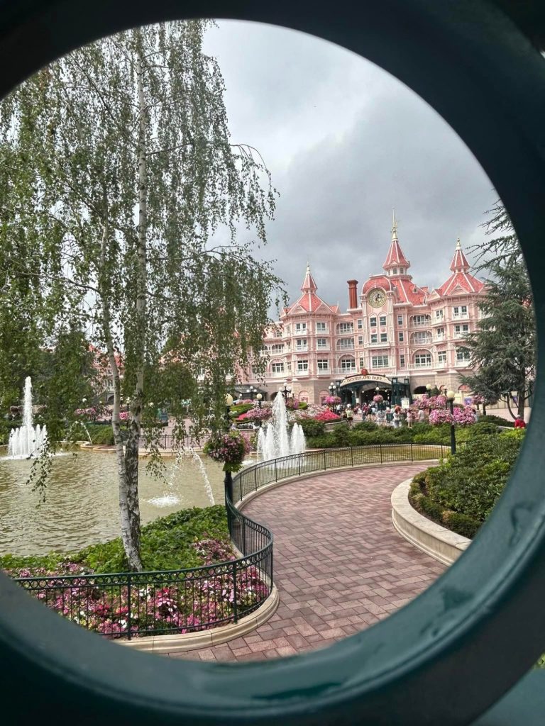 Image of the disneyland hotel taken through a green circular railing, it is a pink victorian style building and there are paths and gardens in front of it