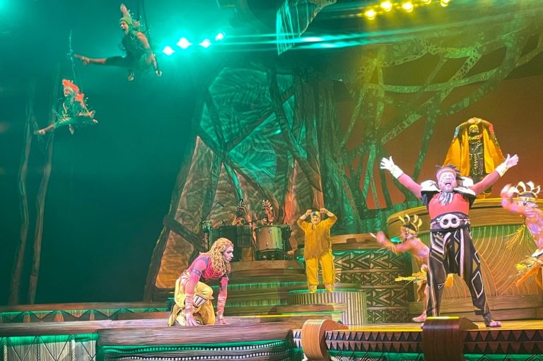 The image shows some of the cast on the drum shaped stage, including Simba kneeling and Pumba with his arms in the air, two acrobats are on ribbons in the air