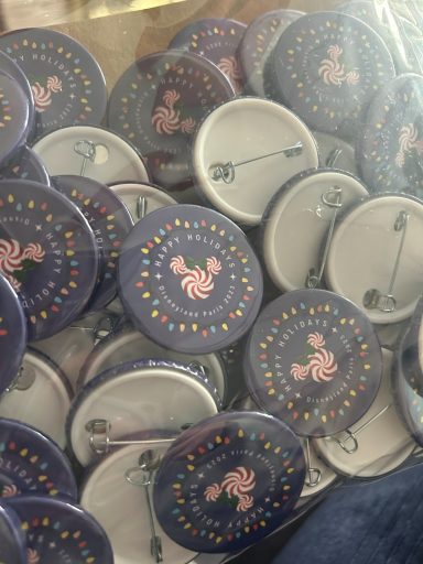 Image of a bag of button badges, all are the same and are purple with a candy swirl shaped mickey head, christmas lights and a message 