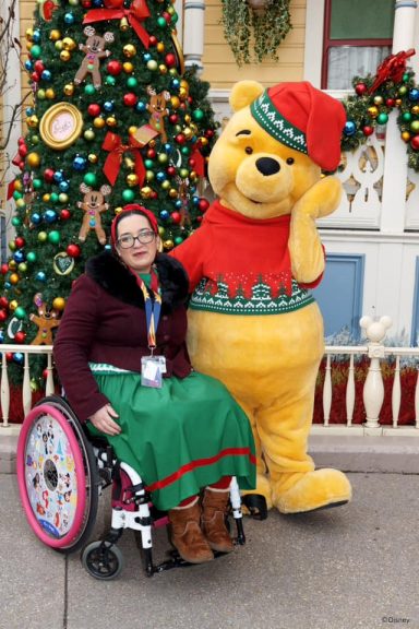 Jody is seated in her chair next to winnie the pooh wearing a Christmas jumper 