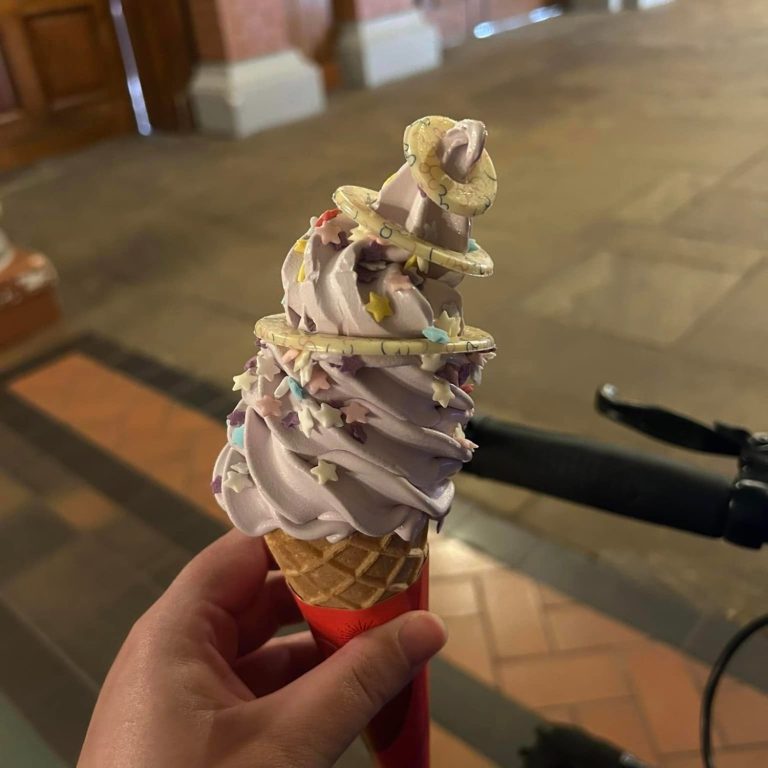 a hand holding a waffle cone filled with a purple soft serve ice cream with sprinkles and white chocolate rings