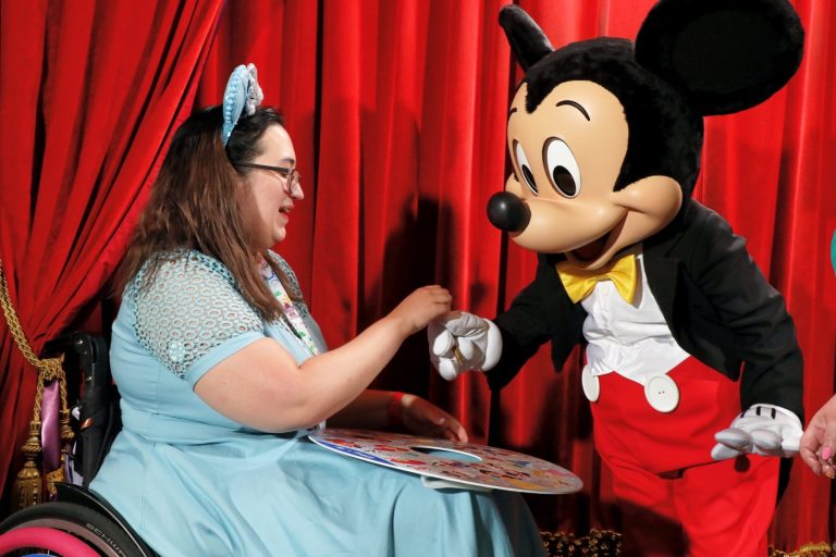 A brunette wearing a blue dress is seated in a wheelchair beside mickey mouse who is taking a pen to sign his autograph