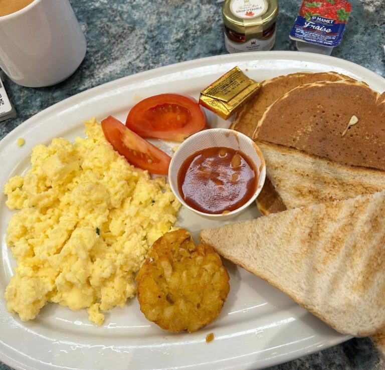 Image shows a plate of breakfast items, toast, pancakes, scrambled egg, tomato, beans and a hash brown