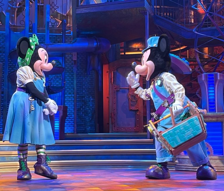 Image shows Mickey and Minnie on stage, wearing blue utility outfits, mickey is carrying a tool box