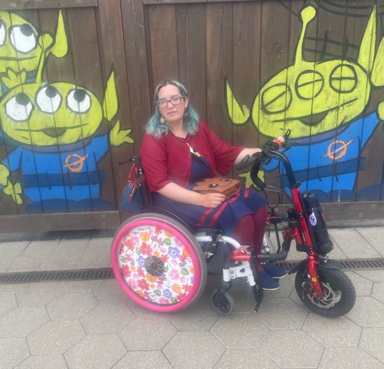 Jody is seated in her chair with the electric handcycle attchment and a gate with paintings of the toy story aliens in the background