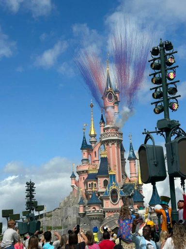 Image shows the dismeyland paris castle in the sunshine with a blue sky and coloured smoke erupting from it