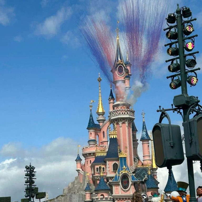 Image shows the disneyland paris castle in the sunshine with daytime fireworks which are colourful smoke coming from it