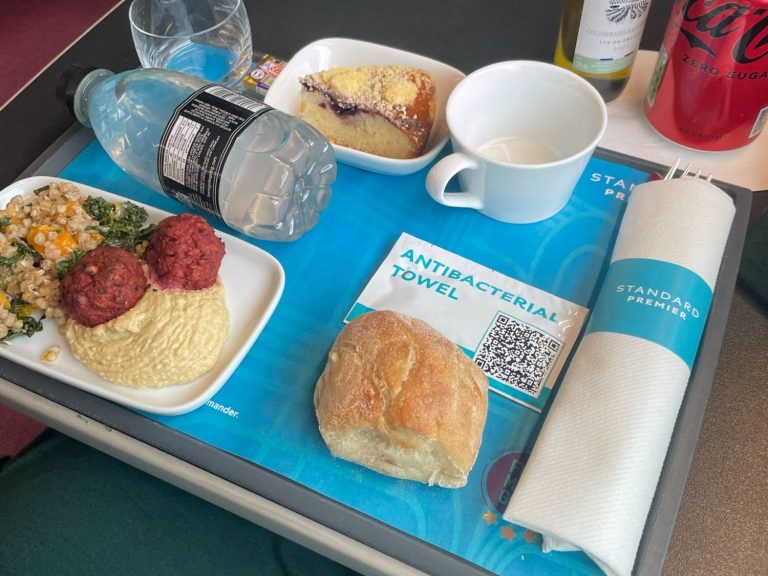 Image showing a tray with a light meal as served on Eurostar, there is a ottle of water, napkin, cup, bread roll, plate of falafals and hummus and a crumble desert